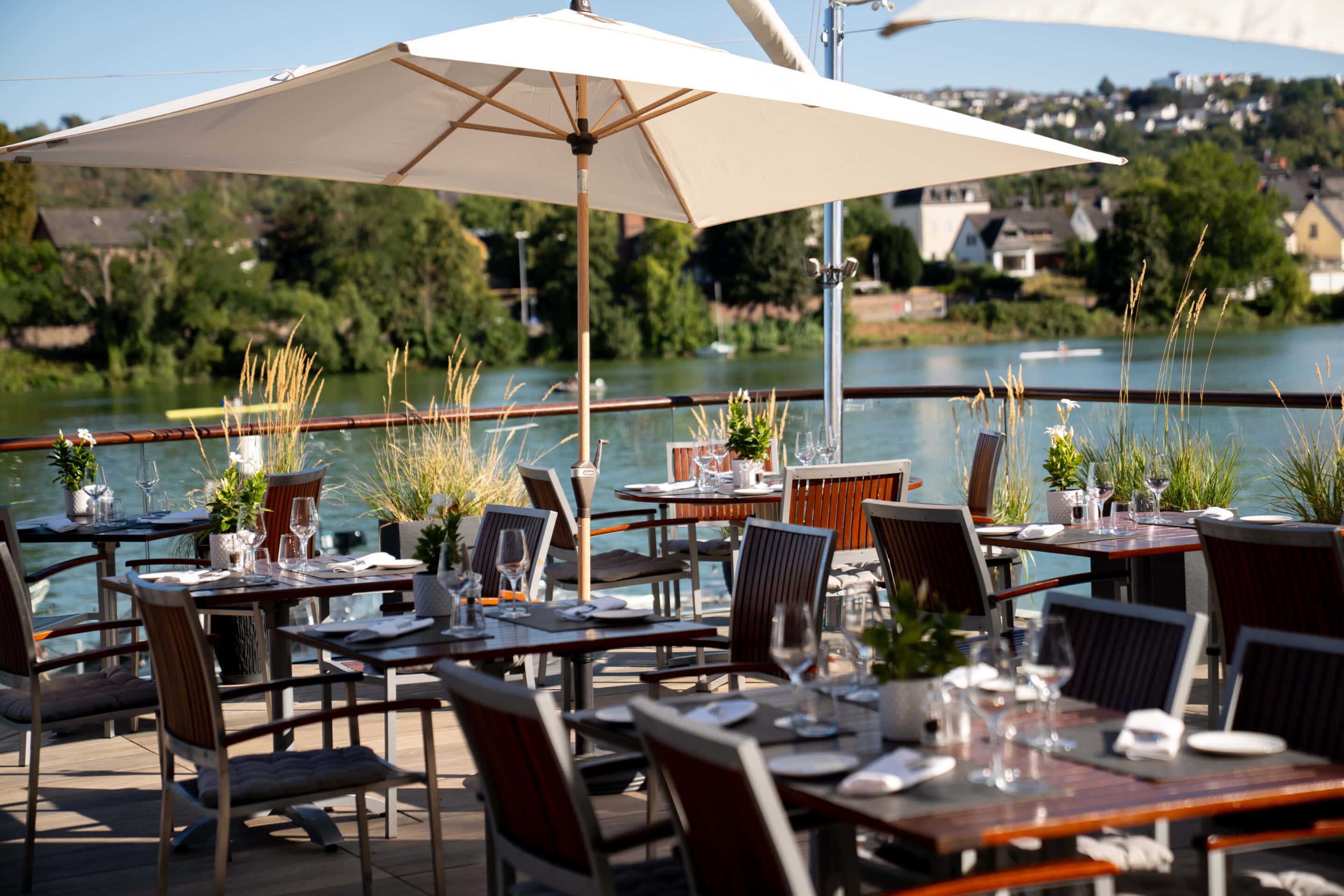 Gedeckte Tische unter weißen Sonnenschirmen auf der Restaurantterrasse des FÄHRHAUS Koblenz mit Blick auf die Mosel.