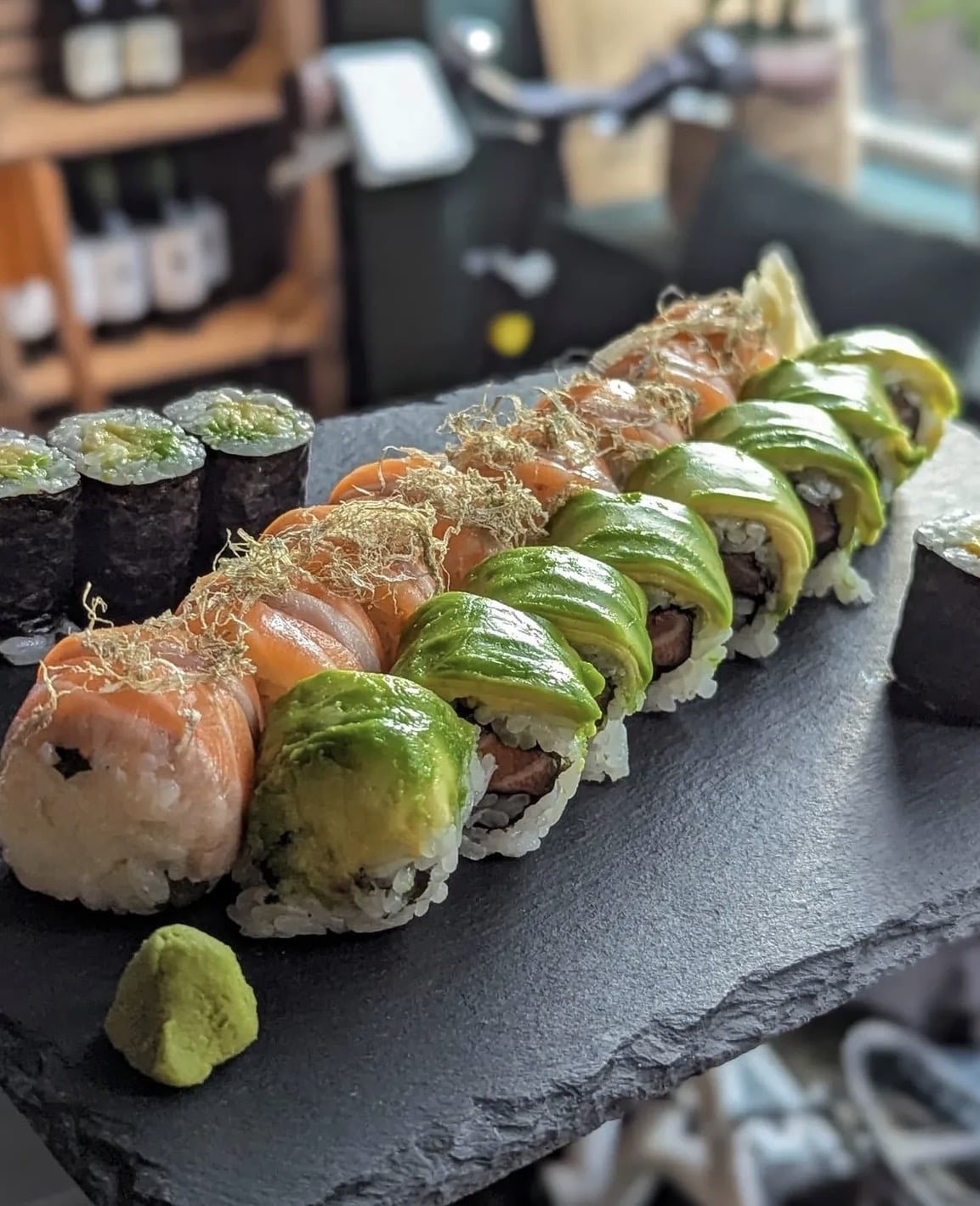 Frisch zubereitete Sushi-Rollen mit Lachs, Avocado und verschiedenen Toppings, angerichtet auf einer schwarzen Schieferplatte mit Wasabi.